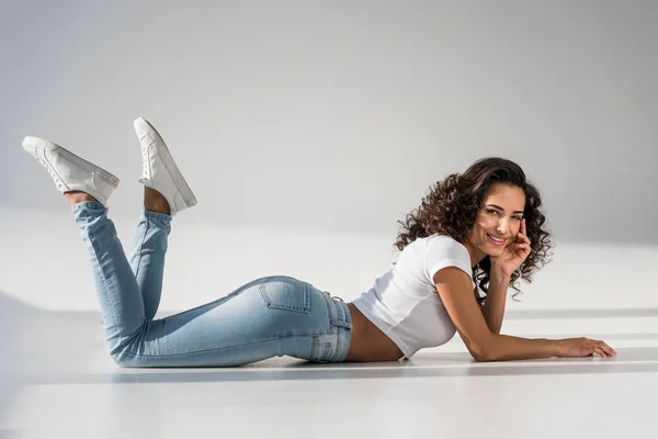 Pretty smiling girl in jeans lying on grey background — Stock Photo