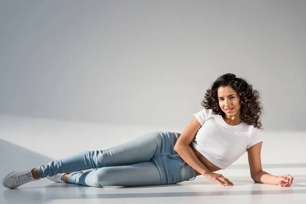 Красивая кудрявая девушка в джинсах лежит на сером фоне — стоковое фото