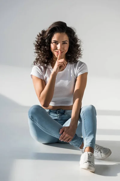 Привлекательная кудрявая девушка в джинсах касается губ пальцем на сером фоне — стоковое фото