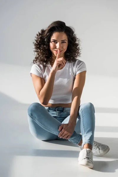 Menina encaracolado atraente em jeans tocando os lábios com o dedo no fundo cinza — Fotografia de Stock