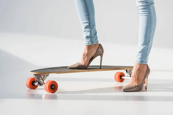 Vista cortada de mulher em jeans e sapatos de salto alto em pé sobre lonboard em fundo cinza — Fotografia de Stock