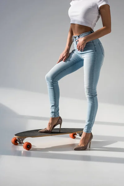 Vista cortada de menina sexy em jeans e sapatos de salto alto em pé no longboard de madeira no fundo cinza — Fotografia de Stock