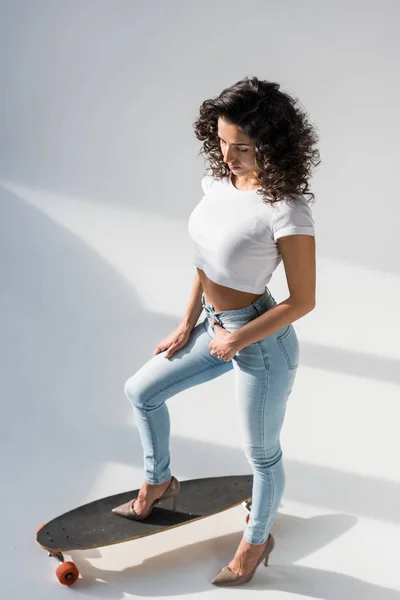 Mulher encaracolada em jeans e top de cultura em pé no longboard em fundo cinza — Fotografia de Stock
