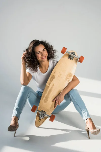 Glamorous girl in high-heeled shoes sitting on floor with longboard on grey background — Stock Photo
