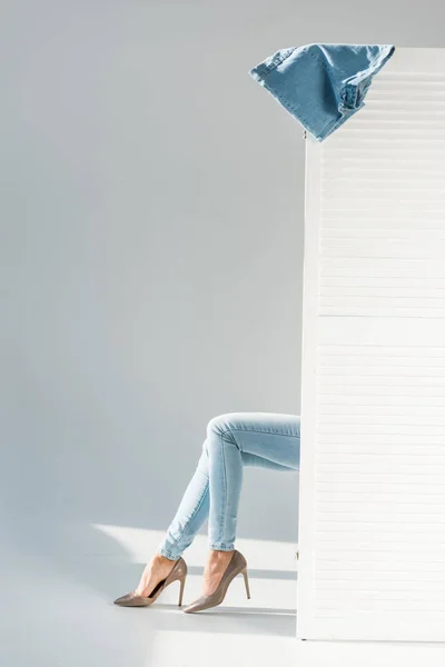 Частковий вигляд жінки в джинсах, що сидять за роздільником кімнати на сірому фоні — стокове фото