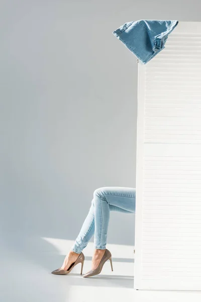 Vista parcial de la mujer en jeans sentada detrás del separador de la habitación sobre fondo gris - foto de stock