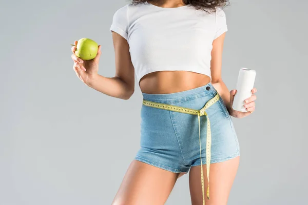 Cropped view of woman in shorts with measuring tape holding green apple and beverage isolated on grey — Stock Photo