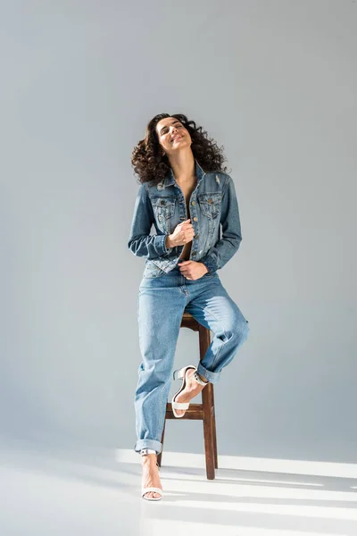 Blissful young woman in jeans and jacket sitting on wooden chair on grey background — Stock Photo