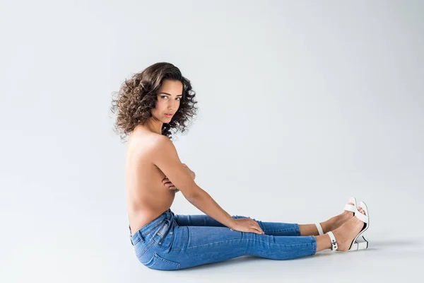 Приголомшлива дівчина в джинсах з оголеними грудьми сидить на сірому фоні — стокове фото