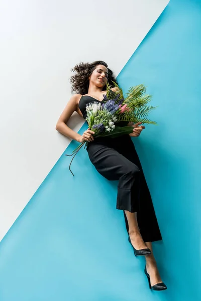 Chica romántica en traje negro con flores tumbadas sobre fondo azul y blanco - foto de stock