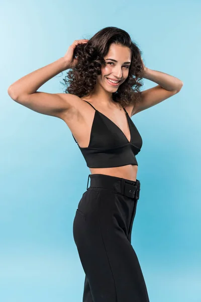 Preciosa mujer sonriente en ropa negra tocando el pelo sobre fondo azul - foto de stock