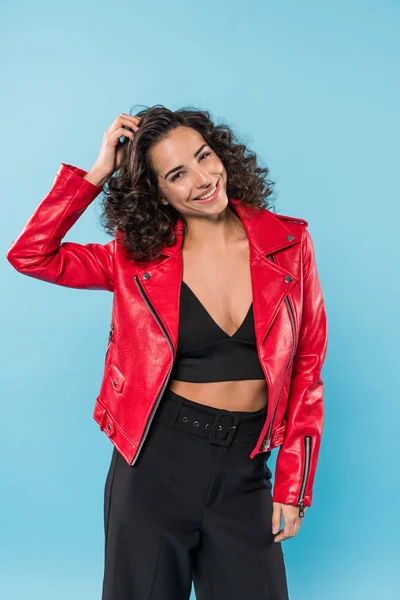 Stylsih girl in red jakcet touching curly hair on blue background — Stock Photo