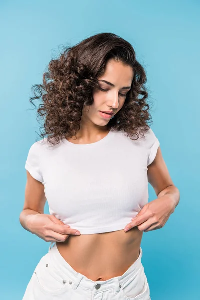 Chica pensativa con el pelo rizado tocando la parte superior de la cosecha en backgorund azul - foto de stock