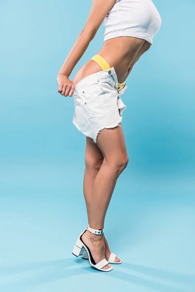 Vista parcial de la mujer elegante en bragas amarillas quitándose pantalones cortos sobre fondo azul - foto de stock