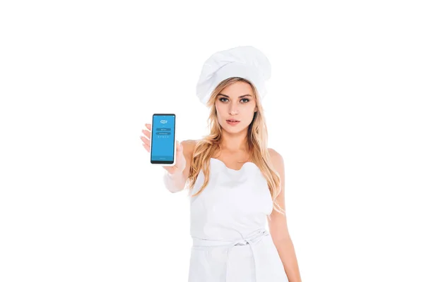 Mulher cozinheira loira atraente em uniforme segurando smartphone com aplicativo skype na tela isolada no branco — Fotografia de Stock