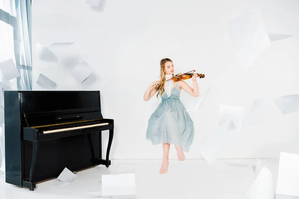 Fille flottante en robe bleue jouant du violon avec des feuilles de papier dans l'air — Photo de stock