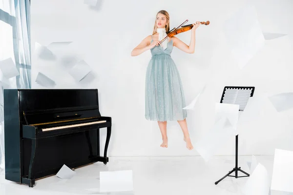 Плавающая девушка в синем платье играет на скрипке возле фортепиано с листами бумаги в воздухе — стоковое фото