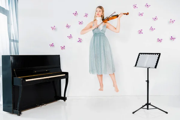 Chica flotante en vestido azul tocando el violín cerca del piano sobre fondo blanco con mariposas - foto de stock