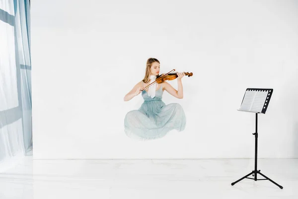 Chica flotante en vestido azul tocando violín con soporte de música sobre fondo blanco - foto de stock