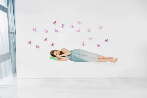 Niña en vestido azul durmiendo en libro en el aire sobre fondo blanco con mariposas púrpura - foto de stock