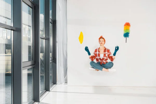 Chica flotante en jeans y camisa a cuadros meditando con trapo y plumero sobre fondo blanco - foto de stock