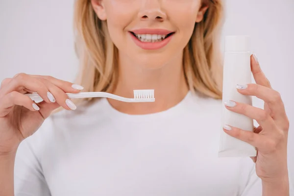 Обрезанный вид улыбающейся женщины, держащей зубную щётку и зубную пасту с выделенным на белом пространством — стоковое фото