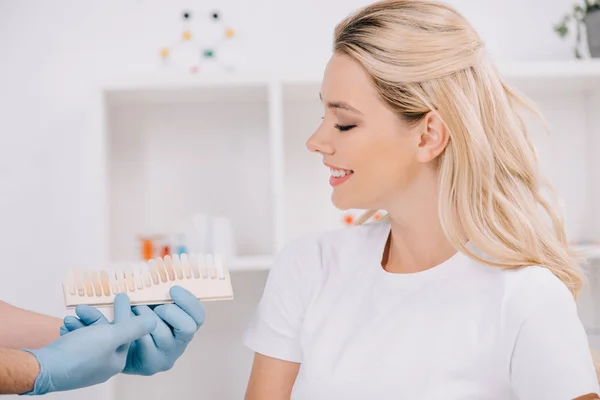 Красива жінка дивиться на стоматолога, який тримає кольорову палітру зубів у клініці — стокове фото