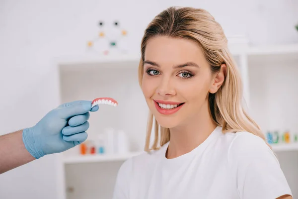 Beautiful smiling woman looking at camera while dentist in latex gloves holding teeth cover in clinic — Stock Photo
