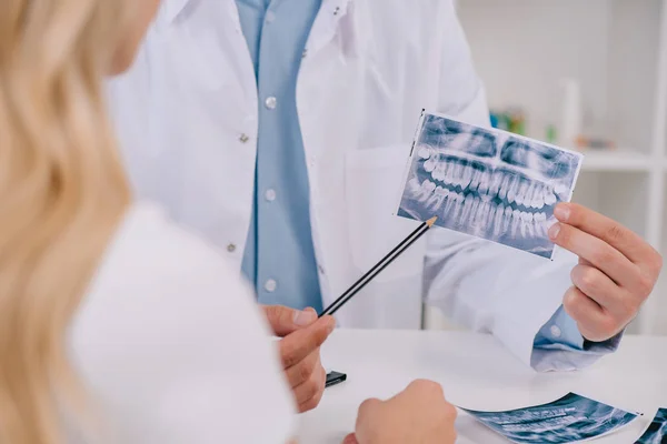 Vista recortada del ortodoncista apuntando a la radiografía de los dientes durante la consulta con la paciente femenina en la clínica - foto de stock