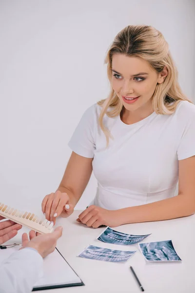 Bella donna sorridente che punta con il dito la tavolozza dei colori dei denti durante la consultazione con il dentista isolato sul bianco — Foto stock