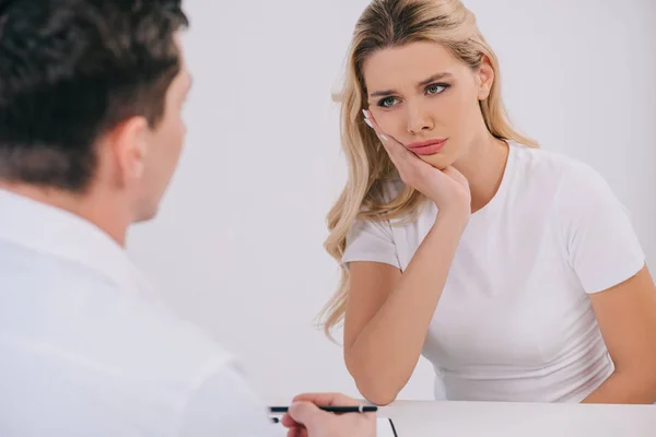 Hermosa mujer tocando la cara y tener dolor de muelas durante la consulta con el dentista masculino aislado en blanco - foto de stock