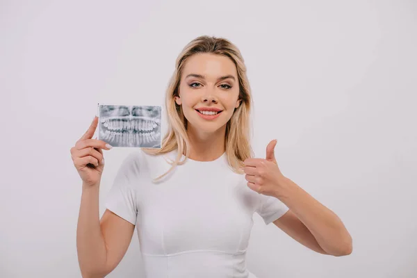 Hermosa mujer en camiseta sosteniendo los dientes de rayos X, mostrando el pulgar hacia arriba y mirando a la cámara aislada en blanco - foto de stock