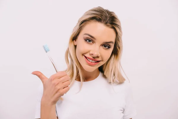 Bela mulher em camisa branca segurando escova de dentes, olhando para a câmera e mostrando sinal de polegar para cima isolado no branco — Stock Photo