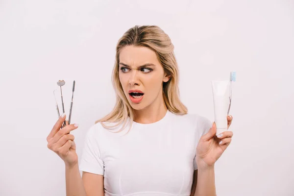 Bela mulher chocada segurando pasta de dentes, escova de dentes e instrumentos dentários isolados em branco — Fotografia de Stock