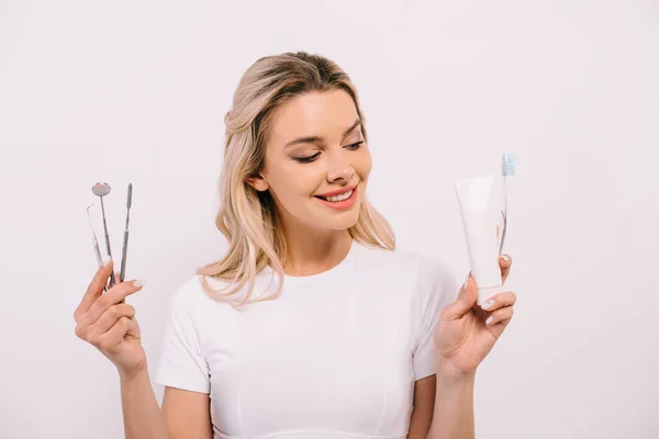 Bela mulher sorridente segurando pasta de dentes, escova de dentes e instrumentos dentários isolados em branco — Fotografia de Stock