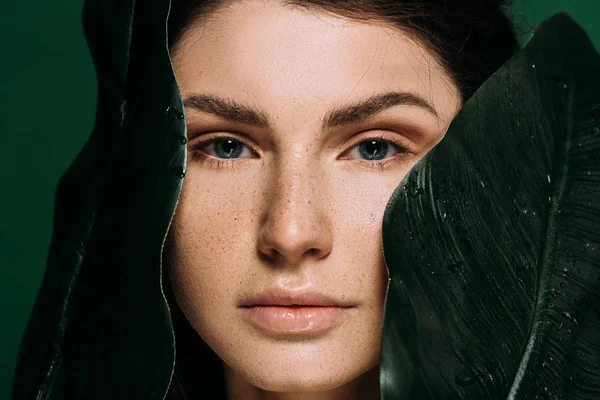 Beautiful girl with freckles on face posing with palm leaves isolated on green — Stock Photo