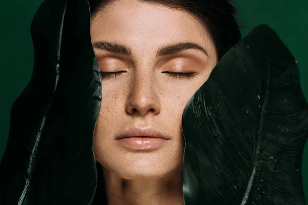 Retrato de hermosa chica con los ojos cerrados posando con hojas verdes aisladas en verde - foto de stock