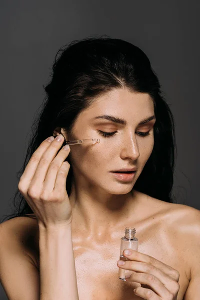 Naked brunette girl applying serum on face with pipette isolated on grey — Stock Photo