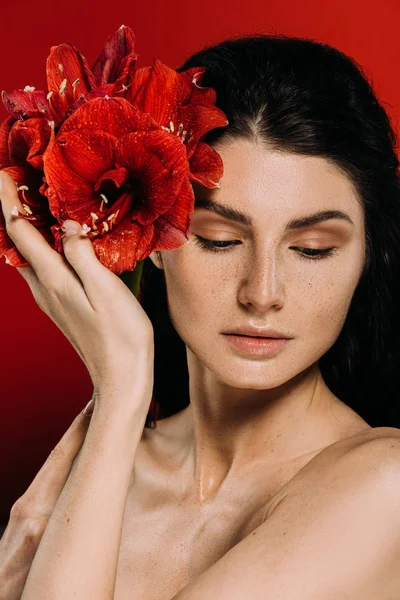 Belle femme avec des taches de rousseur sur le visage posant avec des fleurs d'amaryllis, isolé sur le rouge — Photo de stock