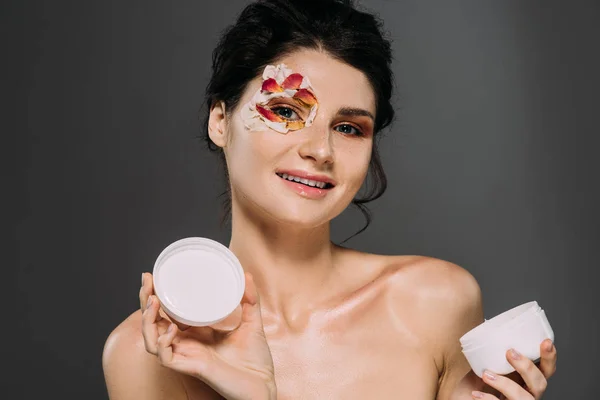 Beautiful woman with petals on face holding plastic container with moisturizing cream isolated on grey — Stock Photo