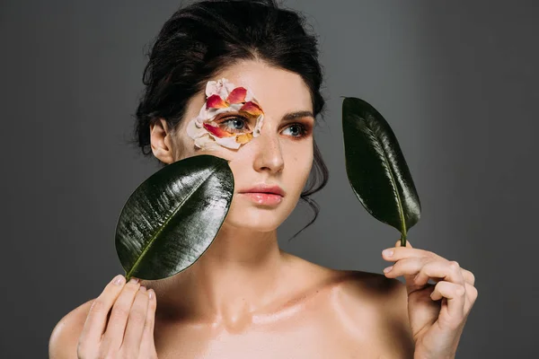 Atractiva mujer con pétalos de flores alrededor del ojo sosteniendo hojas aisladas en gris - foto de stock