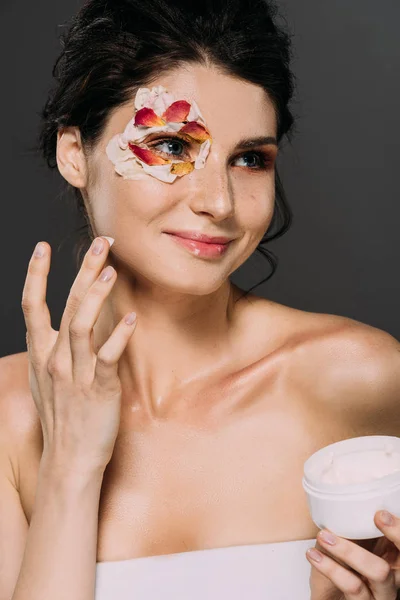 Bela mulher sorridente com pétalas no rosto aplicando creme hidratante isolado no cinza — Fotografia de Stock