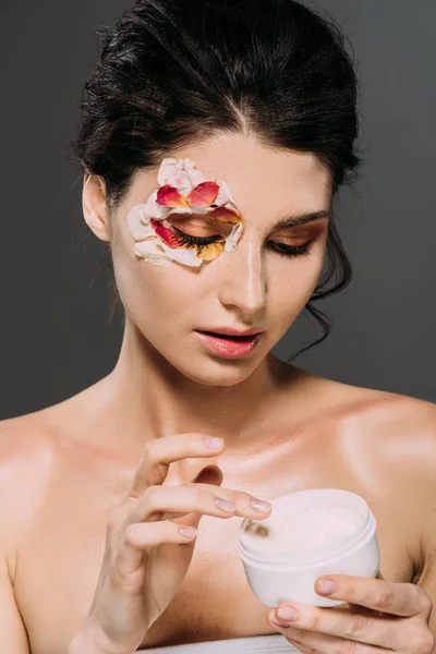 Attractive young woman with petals on face holding plastic container with cream isolated on grey — Stock Photo