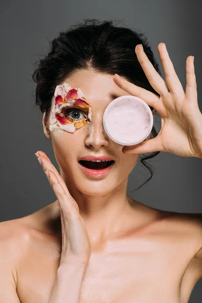 Surprised girl with petals on face holding plastic container with cream isolated on grey — Stock Photo