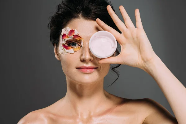 Beautiful brunette girl with petals on face holding cosmetic cream isolated on grey — Stock Photo