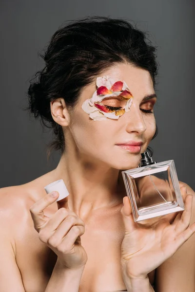 Attractive tender girl with closed eyes and petals on face smelling perfume isolated on grey — Stock Photo