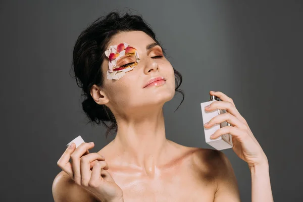 Hermosa chica tierna con los ojos cerrados y pétalos en la cara rociando perfume aislado en gris - foto de stock