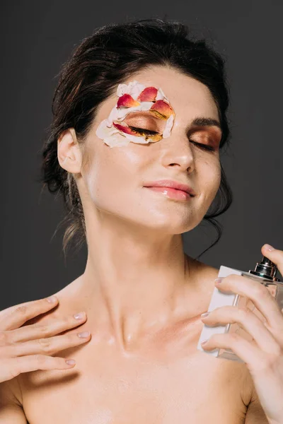 Carefree girl with closed eyes and petals on face spraying perfume isolated on grey — Stock Photo