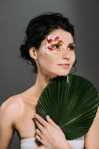 Hermosa mujer de ensueño con pétalos florales alrededor de ojo sosteniendo hoja aislada en gris - foto de stock