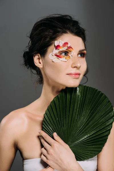 Bella donna sognante con petali floreali intorno alla foglia tenuta d'occhio isolato su grigio — Foto stock